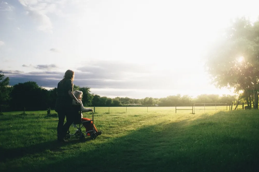 A carer walking with a resident in a wheelchair across a field at sunset